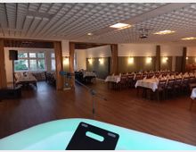 Ein Veranstaltungsraum mit gedeckten Tischen und Holzstühlen, beleuchtet von Wandlampen und natürlichem Licht durch große Fenster. Im Vordergrund ein Mikrofon auf einem Stativ, der Boden besteht aus Holz und die Decke hat ein geometrisches Muster.