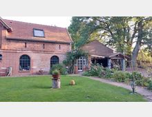 Backstein-Gebäudekomplex mit roten Dachziegeln, bestehend aus einem Hauptgebäude mit großen Rundbogenfenstern und einem angrenzenden Holzcarport. Im Vordergrund eine gepflegte Rasenfläche mit Blumenbeeten und einem gepflasterten Gehweg, umgeben von Bäumen und weiteren Pflanzen.