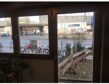 Blick durch ein großes Fenster auf eine städtische Straße mit geparkten Fahrrädern, einer Holzbarrikade und einer gepflasterten Fläche im Vordergrund. Im Hintergrund sind moderne Betongebäude, eine Brücke und bunte Graffiti-Wände zu sehen.