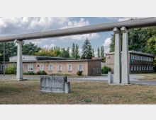 Industriegelände mit flachen, braunen Gebäuden und einer erhöhten Rohrleitung auf Betonstützen im Vordergrund. Die Umgebung ist von Bäumen umgeben, und der Boden besteht aus Rasen mit einzelnen betonierten Bereichen.