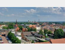 Stadtansicht mit gemischter Bebauung aus Wohnhäusern, einer roten Backsteinkirche mit spitzem Turm sowie Industriegebäuden und Schornsteinen im Hintergrund. Umgeben von Bäumen und parkenden Autos an einem sonnigen Tag.