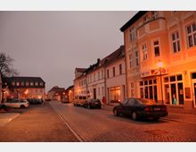 Straßenszene in einer kleinen Stadt am Abend mit gepflasterter Fahrbahn, geparkten Autos und Gebäuden mit historischen Fassaden in warmem Licht, beleuchtet von Straßenlaternen. Im Vordergrund ist ein gelbes Gebäude mit Schaufenstern und einer Beschilderung zu sehen.