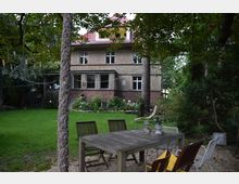 Ein Garten mit Rasenfläche und einem großen Holztisch mit Stühlen im Vordergrund, umgeben von Bäumen und Sträuchern. Im Hintergrund steht ein zweistöckiges Wohnhaus mit Backsteinsockel, verputzter Fassade und einem roten Ziegeldach.