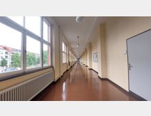Langer Flur in einem Gebäude mit beigen Wänden, glänzendem braunem Boden und großen Fenstern auf der linken Seite, die Blick auf Bäume und Gebäude ermöglichen. Die rechte Seite zeigt Türen, Heizkörper sowie eine Pinnwand an der Wand.