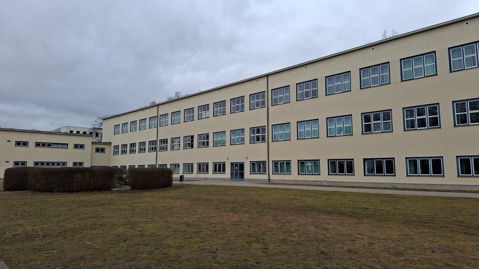 Beiger, mehrstöckiger Schul- oder Verwaltungsbau mit langen Reihen rechteckiger Fensterfronten und flachem Dach. Davor liegt eine große Rasenfläche mit niedrigen Hecken und einem Gehweg unter bewölktem Himmel.