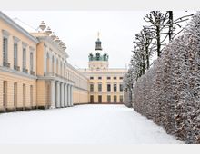 Außenansicht eines barocken Schlosses im Winter, mit einer Fassade in hellgelber Farbe, Säulen und einem Turm mit goldener Statue. Der Hof ist schneebedeckt, und rechts stehen kahle, mit Schnee überzogene Bäume entlang einer Hecke.