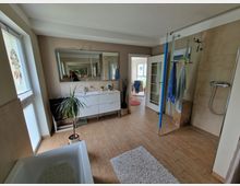 Modernes Badezimmer mit Holzoptik-Fliesenboden, einer freistehenden Badewanne links und einer offenen, bodengleichen Dusche rechts. Ein weißer Waschtisch mit großem Spiegel dominiert die Wand, ergänzt durch eine Zimmerpflanze und Dekorationen; im Hintergrund führt eine Glastür in einen weiteren Raum.