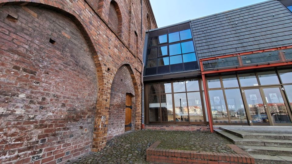 Innenhof mit historischer Backsteinfassade und großen gemauerten Rundbögen, daneben ein moderner Glas-Anbau mit großen Fensterflächen. Kopfsteinpflaster, eine kleine Holz­tür und eine Treppe führen zu einem verglasten Eingangsbereich unter einem schiefergedeckten Dach.