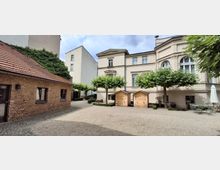 Innenhof zwischen historischen Gebäuden mit heller Putzfassade und großen Rundbogenfenstern sowie einem niedrigen Backsteinhaus mit Ziegeldach. Auf dem geschotterten Platz stehen beschnittene Bäume, zwei kleine Holzhütten und Sitzmöbel mit geschlossenem Sonnenschirm.