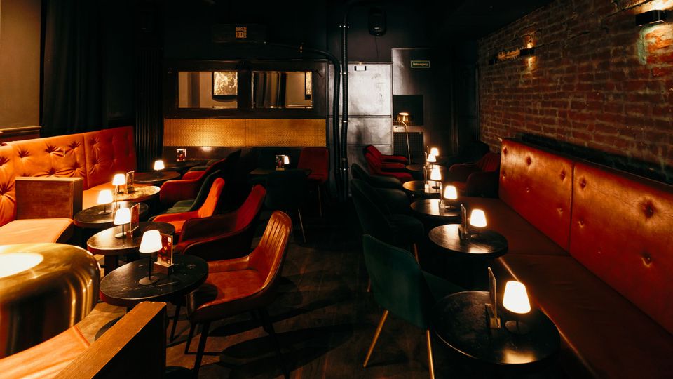 Gemütlicher Bar- oder Lounge-Innenraum mit roten gepolsterten Sitzbänken, kleinen runden Tischen und vielen niedrigen Tischlampen. Dunkle Wände und eine freiliegende Ziegelwand mit Wandleuchten prägen den Raum, dazu stehen Sessel in Reihen vor einer abgetrennten Rückwand.