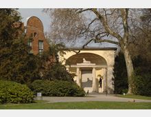 Historisches Gebäude mit gelber Fassade und einem Eingang mit Säulen und einem Reliefbogen darüber, verziert mit einer Löwenfigur. Daneben ein rotes Ziegelhaus, umgeben von Bäumen und Büschen in einer parkähnlichen Umgebung.