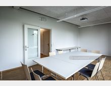 Heller Besprechungsraum mit weißen Wänden, einem Holzboden und einem rechteckigen Tisch in der Mitte, umgeben von Stühlen mit Holzlehnen. Im Hintergrund befinden sich ein Glastür-Eingang sowie weitere Tische an der Wand.