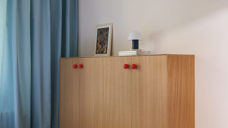 Minimalist bedroom corner with a built-in platform bed featuring under-drawers, topped with a blue duvet. A light wood cabinet with red knobs sits against a pale wall beside floor-to-ceiling blue curtains, with a small lamp and framed artwork on top.