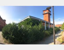 Rotes Backsteingebäude mit einem historischen Turm im Hintergrund, flankiert von moderner Architektur mit weißen Säulen, umgeben von dichter grüner Vegetation und einem gepflasterten Weg im Vordergrund. Der Himmel ist klar und sonnig.