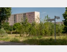 Ein Wohngebietspark mit erhöhtem Gras und jungen Bäumen, im Hintergrund ein Hochhaus mit Balkonen. Rechts steht ein Straßenschild mit den Namen „Raoul-Wallenberg-Straße“ und „Ludwig-Renn-Straße“.