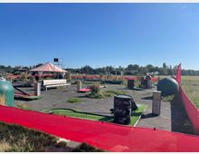 Eine Outdoor-Minigolfanlage mit verschiedenen Bahnen und Hindernissen, umgeben von roten Absperrzäunen. Im Hintergrund steht ein großes Zelt mit rot-weißem Dach, und dahinter sind Bäume und offene Grünflächen zu sehen.