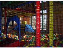 Indoor-Spielbereich mit buntem Bällebad, Klettergerüst aus roten, gelben und blauen Elementen und gesichert durch ein Netz. Im Hintergrund sind Fenster mit Tageslicht erkennbar.