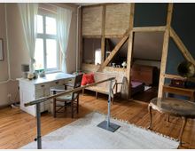 Ein gemütliches Zimmer mit Holzfußboden und sichtbaren Fachwerkbalken. Es ist möbliert mit einem weißen Schreibtisch vor einem Fenster mit weißen Vorhängen, einem Holztisch mit antikem Grammophon, einer Sitzbank und einem Holzbett im Hintergrund. In der Mitte steht eine Haltestange.