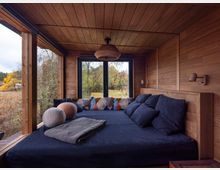 Gemütlicher Raum in einer Holzhütte mit hellen Holzwänden und -decke, bodentiefen Fenstern mit Blick auf eine herbstliche Landschaft. Im Vordergrund befindet sich ein großes, dunkel bezogenes Tagesbett mit bunten Kissen und Decken.