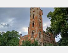 Eine alte, teilweise verfallene Backsteinruine mit einem hohen Turm und mehreren großen Fenstern, umgeben von grünen Bäumen, vor einem bewölkten Himmel.