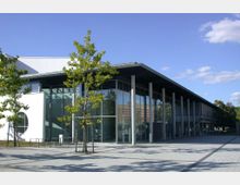 Modernes Gebäude mit großen Glasfassaden und einer Überdachung, die von schlanken Säulen getragen wird. Im Vordergrund befinden sich gepflasterte Wege und junge Bäume, die einen offenen Platz umrahmen.