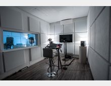 Schallisolierter Aufnahmeraum mit weiß gepolsterten Wänden, einer Glasöffnung zum angrenzenden Raum, einem Mischpult auf einem hohen Tisch, Mikrofonständern und einem Flachbildschirm an der Wand. Ein Lautsprecher steht auf einem Podest, und Kabel verlaufen über den Boden.