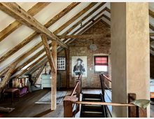 Dachgeschossraum mit freiliegenden Holzbalken, alten Ziegelwänden und Holzfußboden. Ein kleines Fenster mit rotem Rollo, eine hängende Lampe, Möbel wie ein Schreibtisch und eine Kommode sowie eine Treppe in der Mitte dominieren die Szene.