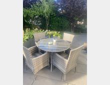 Runde Gartentischgruppe aus grauem Polyrattan mit vier Sesseln auf einer gepflasterten Terrasse. Im Hintergrund stehen große Pflanzkübel mit grünen Pflanzen sowie eine dichte Hecke und Bäume.