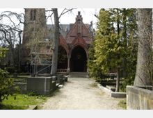 Eine historische Backsteinkirche mit spitzem Giebel und gewölbter Eingangsfront, umgeben von großen Bäumen und Grabsteinen auf einem Friedhof. Vor der Kirche ist ein Gerüst aufgebaut, das auf Renovierungsarbeiten hinweist.