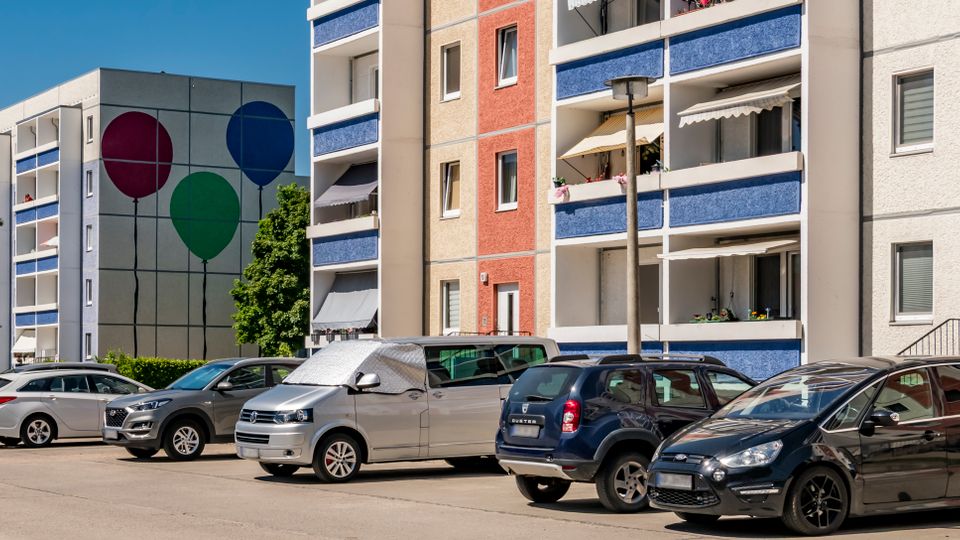 Parkplatz vor mehreren mehrstöckigen Wohnblocks mit Balkonen und farbigen Fassaden in Beige, Rot und Blau. Im Hintergrund ist ein Gebäude mit großer Wandmalerei aus drei Ballons zu sehen, daneben stehen Bäume unter blauem Himmel.