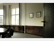 Konferenzraum mit großen Fenstern, Holzvertäfelung an den Wänden und einem langen Tisch mit Stühlen. An der weißen Wand hängen zwei gerahmte Bilder, daneben verlaufen sichtbare Heizungsrohre.