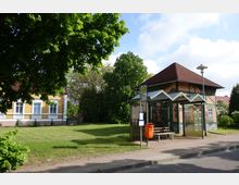 Bushaltestelle mit überdachtem Wartebereich und Sitzbank, flankiert von einer orangefarbenen Mülleimerstation, neben einer Wiese und einem historischen gelben Ziegelgebäude mit weißen Fensterrahmen. Im Hintergrund sind Bäume und ein weiteres Gebäude mit rotem Dach zu sehen.