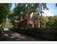 Backsteinhaus mit roten Dachziegeln und grünen Fensterrahmen inmitten einer bewaldeten Umgebung. Im Vordergrund führt ein schattiger Weg entlang eines Zauns, umgeben von Bäumen und Hecken.