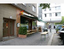 Straßenszene mit einem kleinen Lokal im Erdgeschoss eines Wohngebäudes. Das Lokal hat eine überdachte Terrasse mit Holz- und Metalltischen, bepflanzte Kübel am Rand und ein Werbeschild vor dem Eingang. Im Hintergrund sind andere Gebäude, geparkte Autos und Fußgänger zu sehen.
