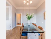 Esszimmer mit einem rechteckigen Glastisch und mehreren Holzstühlen mit blauen Sitzpolstern. Der Raum hat helle Wände, ein Fischgrät-Holzparkett, eine minimalistische Hängelampe und eine Vase mit Blumen auf dem Tisch; im Hintergrund sind eine Tür und ein angrenzender Flur sichtbar.