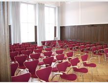 Ein Seminarraum mit einem Parkettboden, bestuhlt mit zahlreichen rot gepolsterten Stühlen auf Metallgestellen. An den Wänden befindet sich eine dunkle Holzvertäfelung, während weiße Vorhänge große Fenster mit hellem Tageslicht einrahmen.