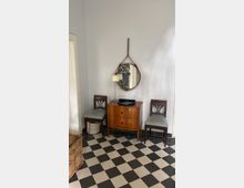 Ecke eines Zimmers mit einem kleinen Holztisch in der Mitte, darauf ein rundes Tablett und eine Lampe. Über dem Tisch hängt ein runder Spiegel mit braunem Lederriemen, flankiert von zwei Holzstühlen mit gestreiften Sitzpolstern. Der Boden ist mit schwarz-weißen Schachbrettfliesen gefliest, links steht ein geflochtener Wäschekorb.