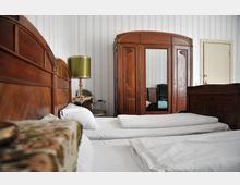 Schlafzimmer mit zwei Einzelbetten, weißen Bettwäsche und einem hölzernen Kopfteil. Im Hintergrund steht ein großer Kleiderschrank mit Spiegel, eine grüne Tischlampe auf einem kleinen Tisch und ein Stuhl. Die Wände sind mit hellen Streifen tapeziert.