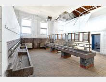 Ehemaliger Waschraum mit mehreren langen, steinernen Waschbecken, veralteten Wasserleitungen und Fliesenboden. Die Decke zeigt Schäden mit offenen Kabeln, und an der Rückseite befinden sich Fenster, die Tageslicht hereinlassen.