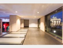 Moderne Innenraumaufnahme mit großen Glasfenstern und Blick auf das beleuchtete Brandenburger Tor bei Nacht. Der Raum hat glatte Steinböden und helle Wände, mit einer Galerieöffnung links, die Kunstwerke zeigt.