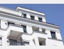 Weiße Fassade eines modernen mehrstöckigen Wohngebäudes mit klaren Linien, großen Fenstern und kleinen Balkonen, umgeben von einem blauen Himmel. Die Architektur weist dezente dekorative Elemente und Geländer mit schlichten Metallmustern auf.