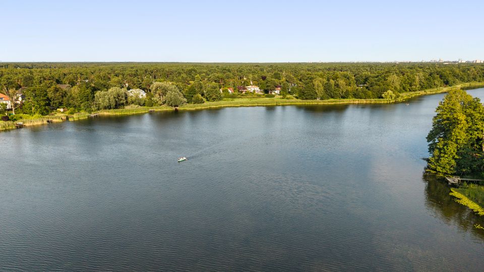 Weitblick über einen breiten See mit ruhiger Wasseroberfläche; am gegenüberliegenden Ufer verläuft ein schmaler Schilfgürtel vor dichtem Wald mit vereinzelten Häusern. Rechts ragt eine bewaldete Landzunge ins Wasser, am Horizont sind in der Ferne Gebäude einer Stadt zu erkennen.