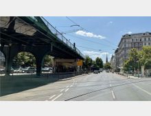 Straßenszene mit einer erhöhten, grünen Eisenbahnbrücke auf der linken Seite, unter der eine Bahnhaltestelle mit wartenden Personen liegt. Rechts verläuft eine breite Straße mit Bäumen und historischen Gebäuden, im Hintergrund ist der Turm einer Kirche zu sehen.