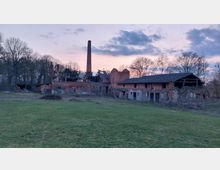 Ruine eines alten Backsteinindustrigebäudes mit zerstörtem Dach, einem hohen Schornstein und umgeben von einem umzäunten grasbewachsenen Gelände. Im Hintergrund stehen Bäume vor einem Himmel in der Abenddämmerung.