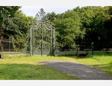 Ein kunstvoll verziertes grünes Metalltor steht am Eingang eines Parks, umgeben von einer halbrunden Gitterzaunanlage. Im Hintergrund sind dichte, grüne Bäume und ein asphaltierter Weg zu sehen.