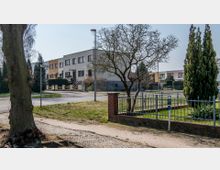 Wohnsiedlung in einer ruhigen Straße mit mehrstöckigen Wohnhäusern, Vorgärten und Gehwegen. Im Vordergrund sind ein Baum, ein Metallzaun und ein Bürgersteig zu sehen; im Hintergrund stehen vereinzelt Bäume und Laternen.