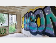 Innenansicht eines verlassenen Gebäudes mit abblätternder Wandfarbe, großen Graffiti an den Wänden und einem zerbrochenen Fenster, das den Blick auf ein überwuchertes Außengelände und ein weiteres verfallenes Gebäude zeigt.