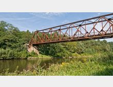 Eisenbahnbrücke aus rostfarbenem Stahl mit einer offenen Fachwerkkonstruktion, die über einen Fluss in einer grünen, bewaldeten Umgebung führt. Im Vordergrund sind Ufervegetation und Gräser zu sehen, während der klare Himmel das Bild ergänzt.