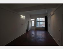 Ein leerer Raum mit weißen Wänden, dunkelrotem Betonboden und einer Fensterfront auf der Rückseite, bestehend aus einer Tür mit Glaseinsatz und mehreren Fenstern. An den Wänden hängen schlichte Leuchtstofflampen, und der Raum hat eine minimalistische Einrichtung.