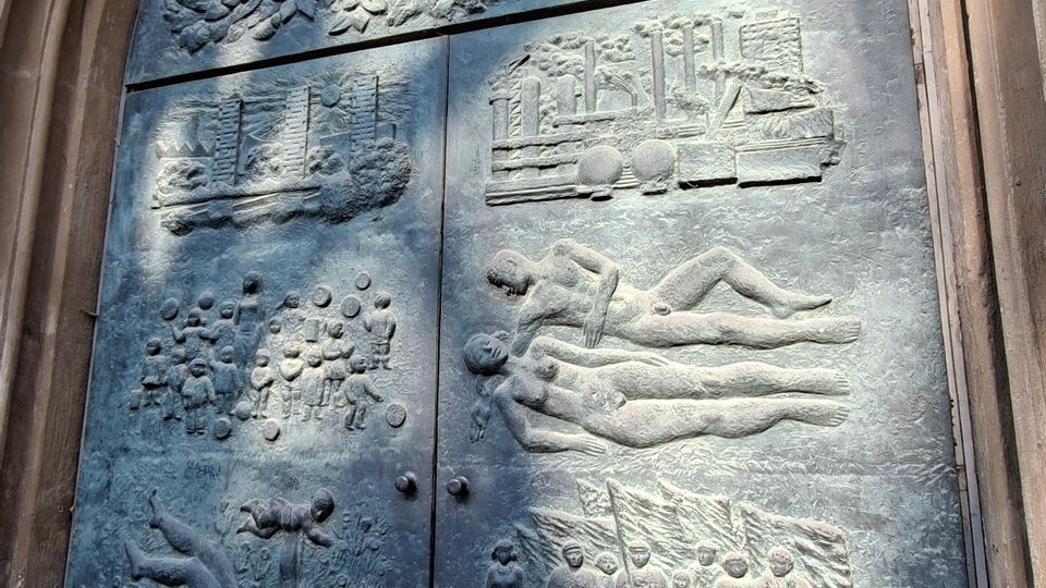 Große bronzene Doppeltür in einem hohen, steinernen Spitzbogenportal, mit reliefartigen Szenen und Figuren auf den Türflügeln. Im oberen Bereich ist ein Baumrelief zu sehen; die Oberfläche wirkt patiniert und wird von schrägem Sonnenlicht teilweise beleuchtet.